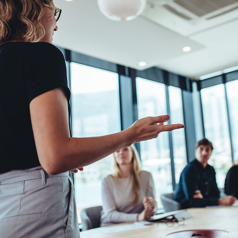 View from the back of a woman holding a presentation to her colleagues in an office.